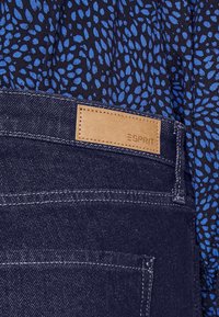 Jeans de mezclilla oscura con una etiqueta de cuero que presenta "ESPRIT". El fondo tiene una tela azul marino con un patrón de hojas azules. Textura suave y costuras resistentes.