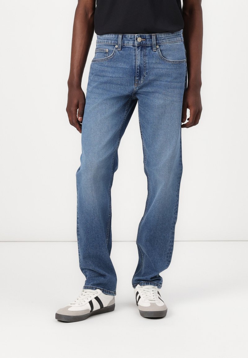 Jeans din denim de un albastru deschis, cu un design drept, stilizare cu cinci buzunare și detalii de cusătură subtile. Purtați cu adidași gri și albi.