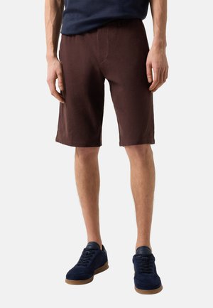 Mann trägt dunkelbraune knielange Shorts, marineblaue Sneaker mit Gummisohlen und ein marineblaues Oberteil, steht vor einem schlichten Hintergrund.
