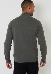 Hombre de espaldas vistiendo un suéter de manga larga gris oscuro y pantalones negros frente a un fondo liso gris claro.