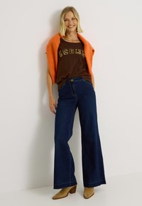 Camiseta marrón con "ICONS" en estampado de leopardo, combinada con unos jeans de pierna ancha de cintura alta en azul oscuro y un cárdigan corto coral. Botas de tacón color beige.