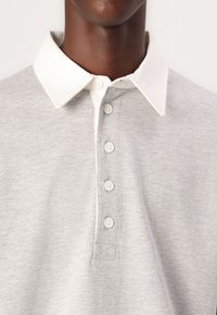 Grå polo skjorte med hvit krage, fem knappelukking, og en myk tekstur. Har synlige sømmer og en brodert detalj på kragen.