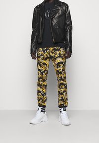 Schwarze Leder-Bikerjacke über einem schwarzen T-Shirt, kombiniert mit gemusterten schwarzen und goldenen Hosen und weißen Sneakern mit schwarzen Akzenten.