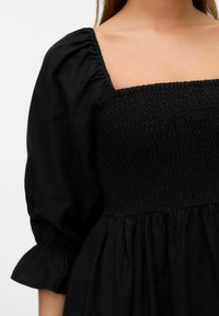 Robe noire avec un décolleté carré, présentant un bodice smocké et des manches bouffantes. Le tissu est lisse avec une jupe fluide et froncée.