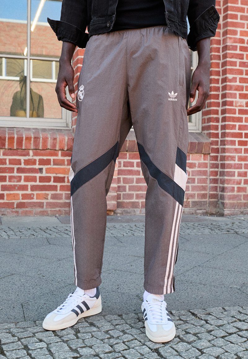 Pantalones deportivos grises con rayas diagonales en negro y blanco, que presentan los logos de Adidas y Real Madrid. Combinados con zapatillas blancas.
