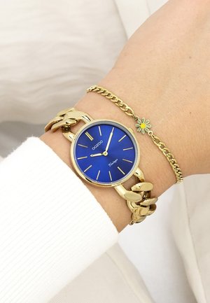 Montre en or avec un cadran bleu, large bracelet en maillons et accents dorés. Bracelet fin en or avec un petit charme en forme de fleur.