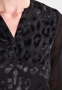 Haut noir avec un design texturé à imprimé léopard et un léger éclat ; manches transparentes avec un léger froncé au niveau de l'encolure.