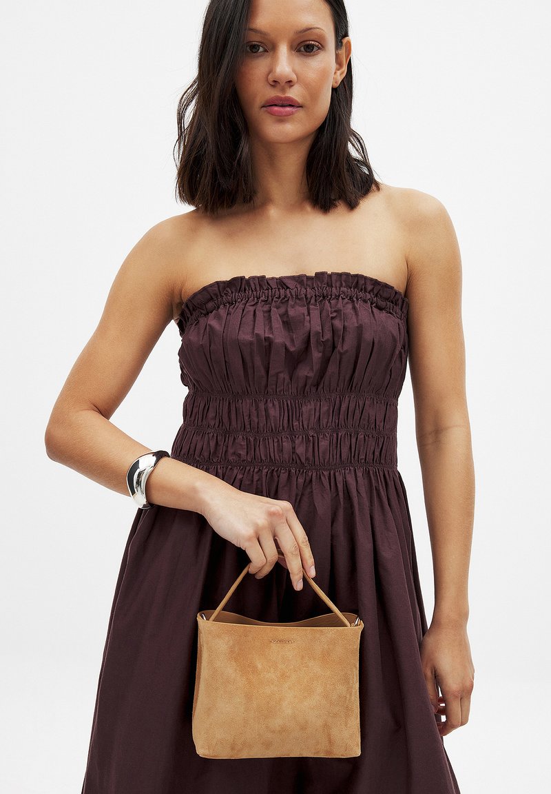 Borsa in suede color tan con forma strutturata e manico superiore, tenuta in mano, abbinata a un vestito senza spalline marrone scuro con ruches.