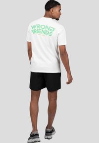 Bílá bavlněná tričko s nápisem "WRONG? FRIENDS" v zeleném grafickém písmu na zadní straně, spárované s černými šortkami a šedými sportovními botami.