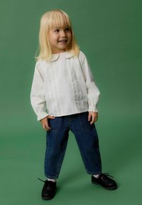 Glunderend jong kind met blond haar staat tegen een groene achtergrond, gekleed in een witte blouse met lange mouwen, blauwe jeans, witte sokken en zwarte schoenen.