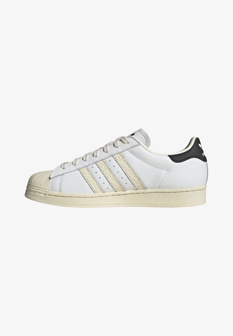 Baltas ādas sneakers ar melnu papēža akcentu, trim bežām svītrām, teksturētu purngalu un gaišas krāsas gumijas zoli, ar perforācijām gaisa caurlaidībai.