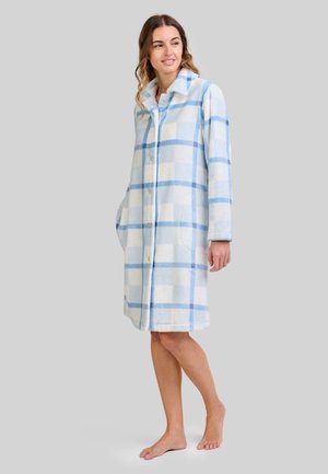 Mujer de pie descalza en un abrigo de forro polar suave, de longitud hasta la rodilla, con un patrón de cuadros azules y blancos, manos en los bolsillos, mirando hacia un lado.