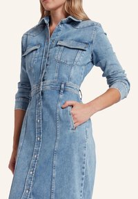 Jeansowa sukienka koszulowa w jasnoniebieskim kolorze, z dwiema kieszeniami na piersiach, zapinana na guziki z przodu, z zamkniętymi mankietami i dopasowanym talią o subtelnej fakturze.