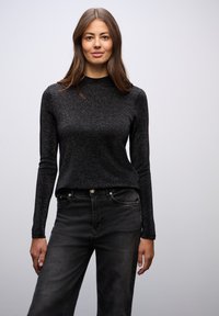 Mustat pitkät hihat -paita, jossa on hillitty kimalteleva pinta, korkea kaula-aukko ja muotoiltu malli, yhdistettynä mustiin farkkuihin, joissa on etutaskut.