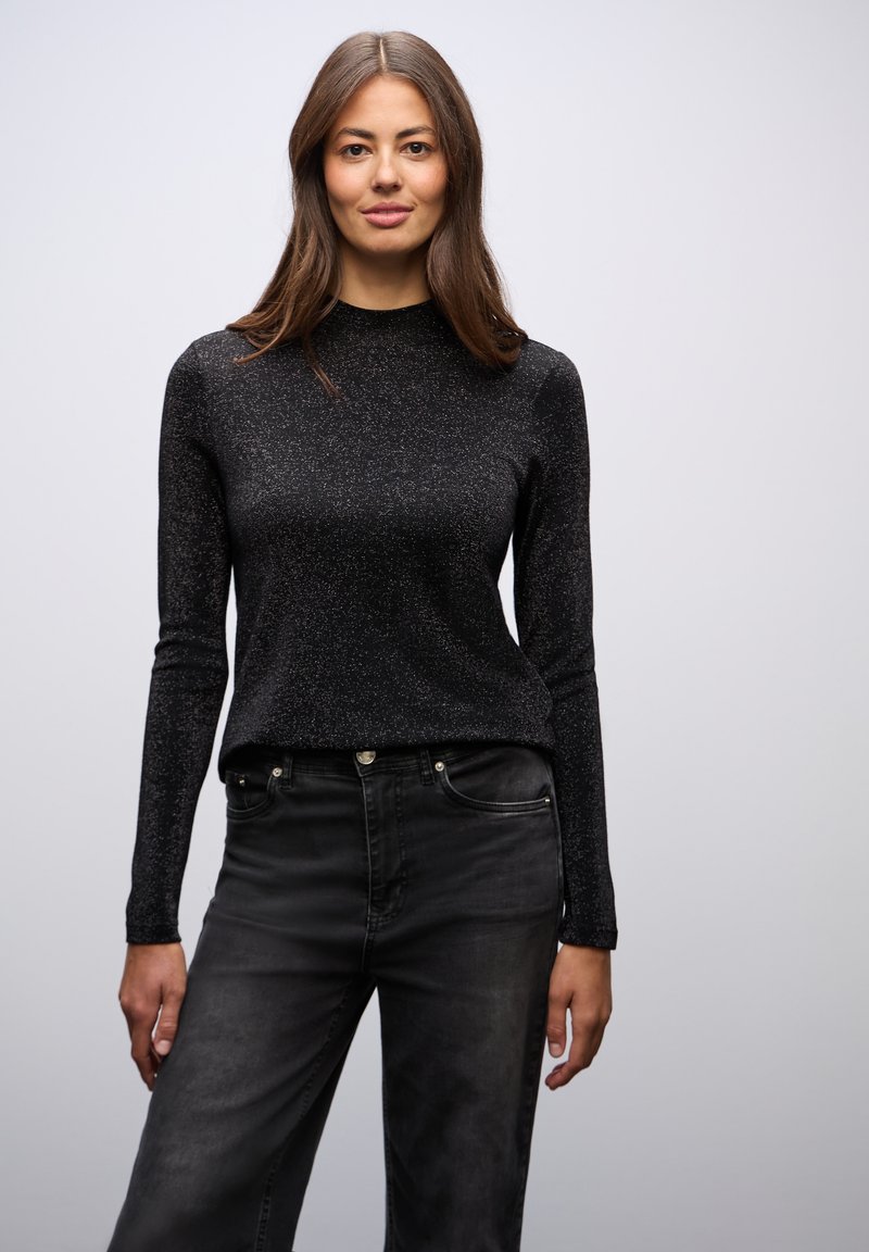 Mustat pitkät hihat -paita, jossa on hillitty kimalteleva pinta, korkea kaula-aukko ja muotoiltu malli, yhdistettynä mustiin farkkuihin, joissa on etutaskut.