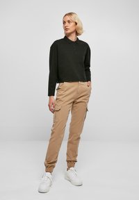 Svart langermet polo-skjorte med krage; beige cargo-bukser med elastiske detaljer og sidelommer; hvite sneakers; minimalistisk bakgrunn.
