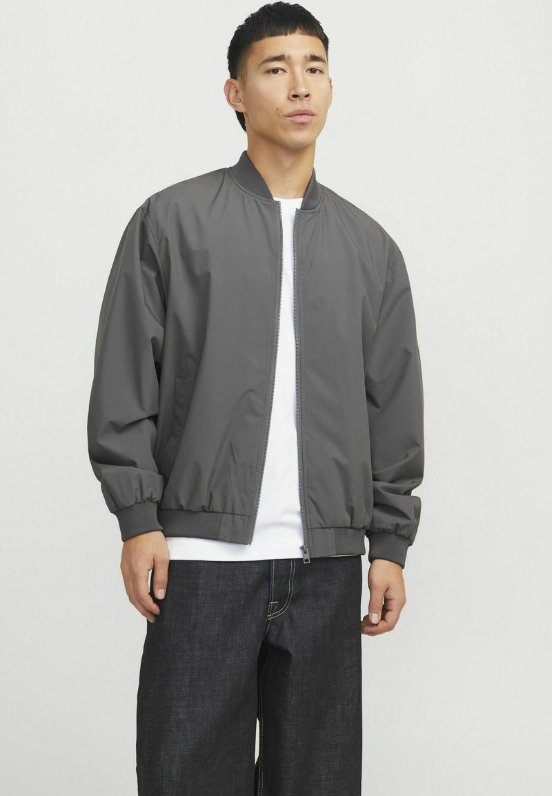 Gray bomber jacket with a front zipper, ribbed cuffs, and waistband, worn over a white t-shirt, paired with dark denim jeans.