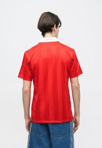 Νεαρός με κοντά σκούρα μαλλιά που φοράει κόκκινο κοντομάνικο πουκάμισο με λευκό γιακά και μπλε τζιν παντελόνι, γυρισμένος με την πλάτη στην κάμερα.