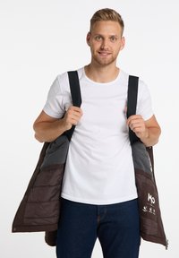 Giacca imbottita marrone con interno grigio, dotata di spallacci neri e chiusura con zip. Indossata su una t-shirt bianca e pantaloni in denim.