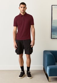 Bordo spalvos polo marškinėliai su užtrauktuku ant apykaklės, juodos kelnaitės su kišenėmis, juodos kojinės ir juodi sportiniai bateliai. Plokščiai megzta apykaklės detalė. Trumpomis rankovėmis ir laisvo kirpimo.