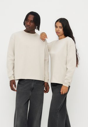 To personer iført beige langærmede toppe og sorte jeans med vidde ben står foran en ensfarvet hvid baggrund.
