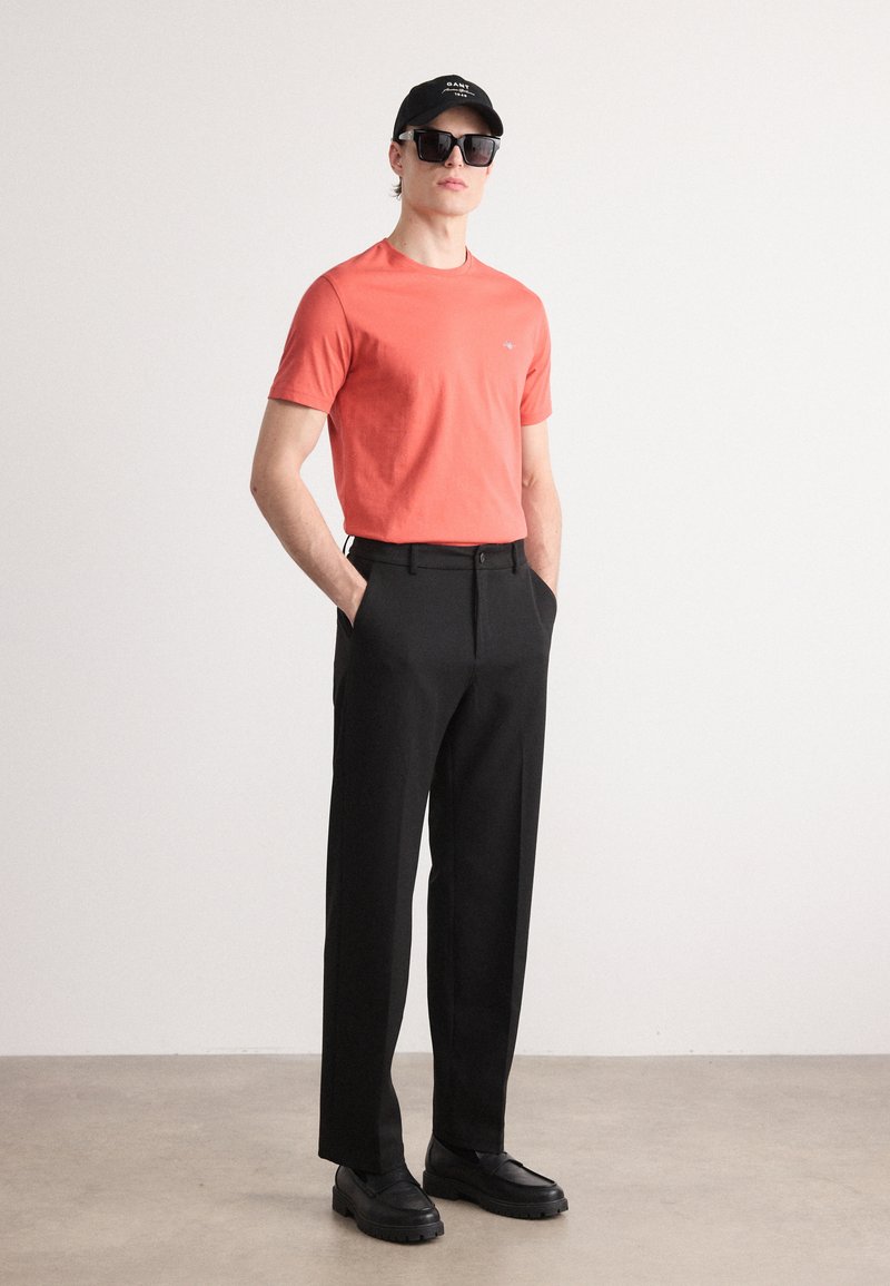 Korallfarbendes Baumwoll-T-Shirt, schwarze maßgeschneiderte Hose, schwarze Mütze, Sonnenbrille und klobige schwarze Schuhe. Schlichtes Design, taillierte Form, klare Linien.