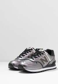 Stříbrné metalické tenisky New Balance s černými detaily, strukturovaným materiálem a bílou mezipodešví. Mají černé tkaničky a logo.
