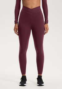 Leggings în nuanță de mahon, cu talie înaltă și un design de talie încrucișat, fabricate dintr-un material neted și elastic, asortate cu încălțăminte sport neagră.