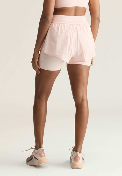 Person trägt hellrosa geschichtete Sportshorts und Sneakers, steht mit leicht gespreizten Beinen mit dem Rücken zur Kamera vor einem einfarbigen Hintergrund.
