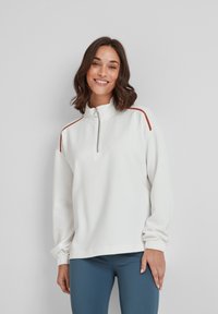 Woman smiling, wearing white zip-up sweatshirt with red stripes on shoulders and blue leggings, standing against a plain light wall.