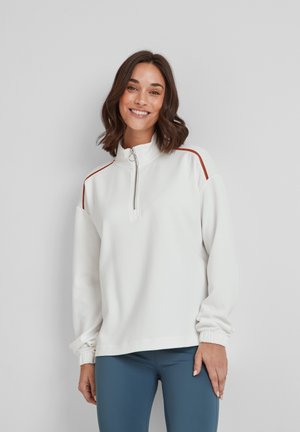 Woman smiling, wearing white zip-up sweatshirt with red stripes on shoulders and blue leggings, standing against a plain light wall.