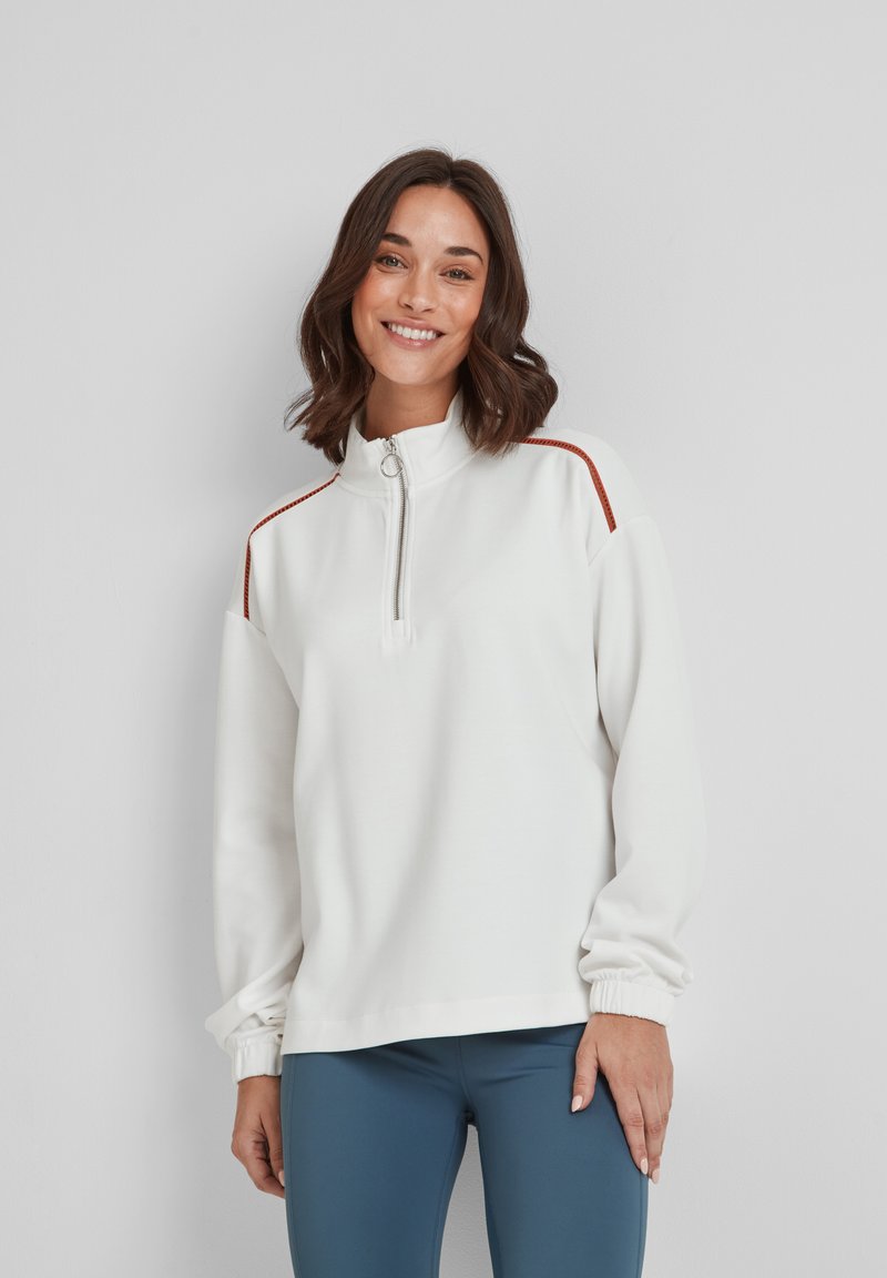 Woman smiling, wearing white zip-up sweatshirt with red stripes on shoulders and blue leggings, standing against a plain light wall.