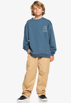 Sudadera azul con cuello redondo y estampado gráfico en el pecho, combinada con pantalones beige de corte holgado y zapatos negros.