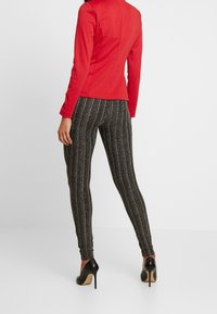 Röd figursydd jacka med långa ärmar, kombinerad med svarta och silverrandiga leggings. Modellen bär svarta högklackade skor. Enfärgad bakgrund.