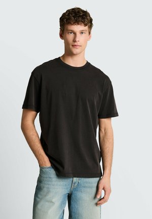 Młody mężczyzna z kręconymi włosami, ubrany w luźny czarny T-shirt i jasnoniebieskie dżinsy, stoi z jedną ręką w kieszeni na jednolitym tle.