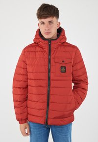 Giacca a vento rossa con design trapuntato orizzontale, chiusura con zip e cappuccio fissato. Presenta una tasca frontale con chiusura a scatto e logo.