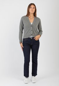 Cardigan grigio a maglia con scollo a V e chiusura con bottoni, abbinato a jeans blu scuro e sneakers bianche.