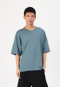 Laisvas, trumpomis rankovėmis t-shirt melsvai žalia medvilnė, su apvalia iškirpte ir nugara sumažintais siūlų kraštais, derinamas su juodomis kelnėmis.