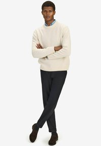 Crème gebreide trui, donkere broek en donkere loafers. De trui heeft een ronde halslijn en ribbelmanchetten. Het model staat met de armen gekruist.
