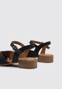 Sandalias de cuero negro con base marrón, correa ajustable con hebilla plateada y tacón de bloque de madera texturizada. Diseño abierto en la parte posterior.