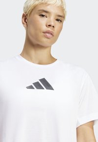 Bijela pamučna majica s crnim Adidas logotipom koji ima tri dijagonalne pruge na prsima. Kratki rukavi i okrugli izrez.