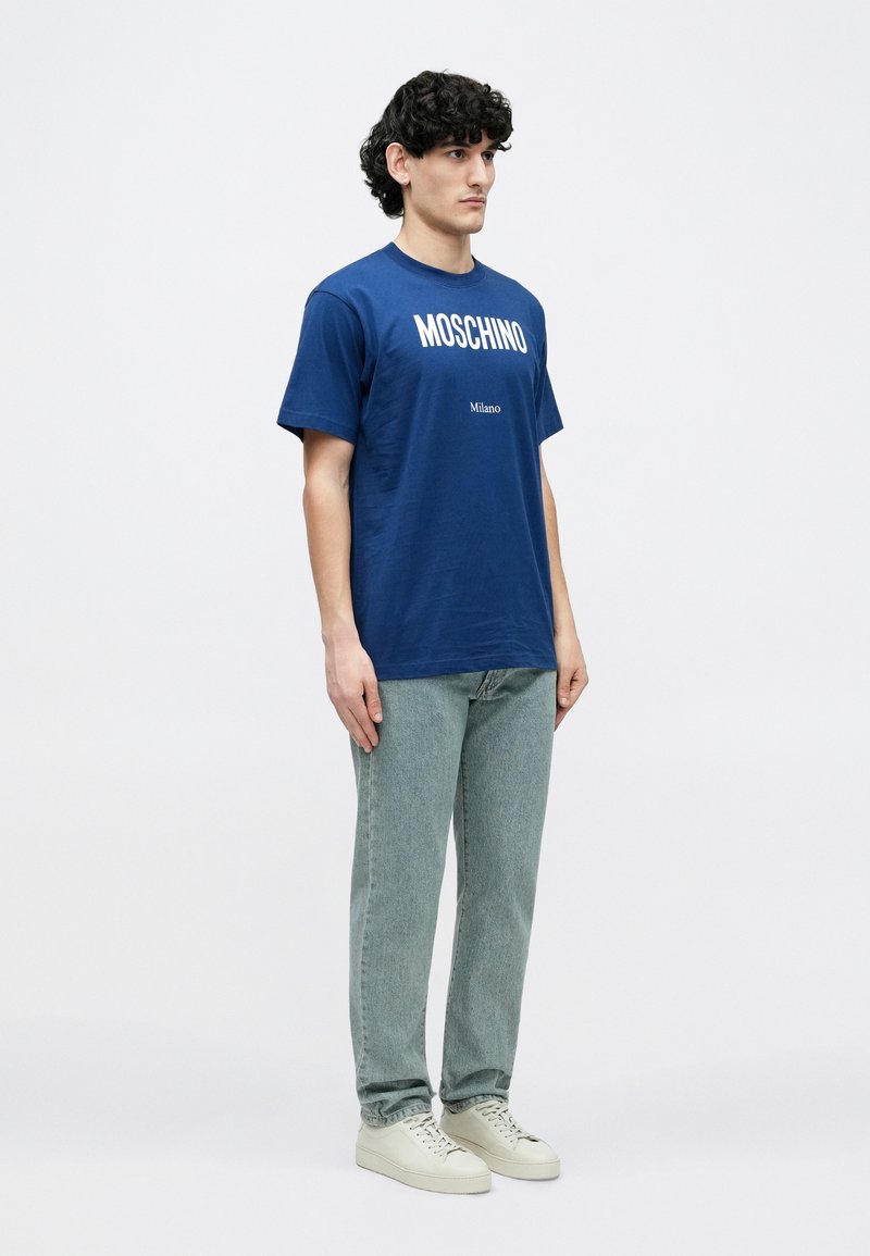 Marineblauw katoenen T-shirt met witte "MOSCHINO"-logo en de tekst "Milano". Model draagt lichtblauwe jeans met rechte pijpen en witte sneakers.