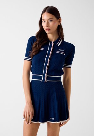 Vrouw met lang bruin haar, gekleed in een marineblauwe gebreide polo-jurk met witte en roze afwerking, staand tegen een effen witte achtergrond.