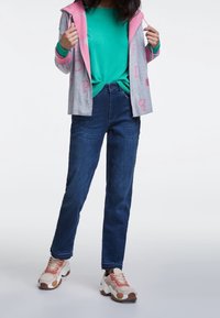Femme portant un pull teal, un sweat à capuche gris avec des lettres roses, un jean bleu foncé et des baskets beiges avec des accents roses, se tenant sur un fond uni.