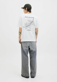 Hvid t-shirt med sort stregtegning af løv og teksten "JUAN-LES-PINS" på bagsiden; parret med løse, lysegrå jeans og sorte støvler.