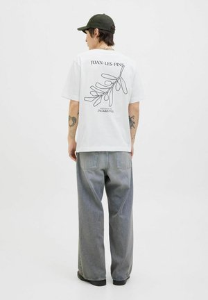 Hvid t-shirt med sort stregtegning af løv og teksten "JUAN-LES-PINS" på bagsiden; parret med løse, lysegrå jeans og sorte støvler.
