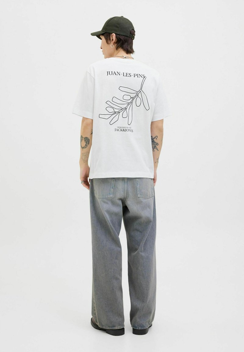 Hvid t-shirt med sort stregtegning af løv og teksten "JUAN-LES-PINS" på bagsiden; parret med løse, lysegrå jeans og sorte støvler.