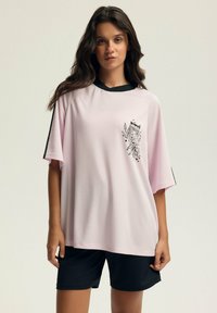 Jasnoróżowa, krótko rękawowa koszulka sportowa z siateczki z czarnymi akcentami i graficznym nadrukiem. Połączona z czarnymi szortami sportowymi.