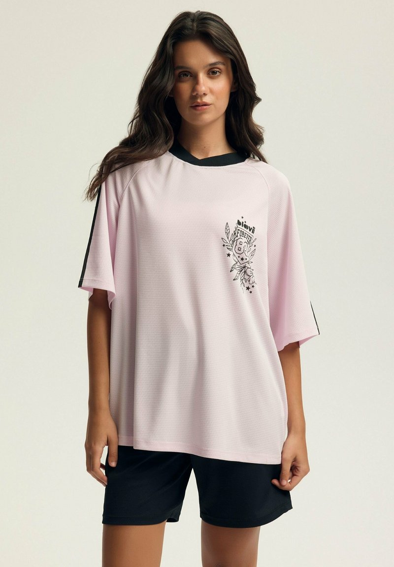 Jasnoróżowa, krótko rękawowa koszulka sportowa z siateczki z czarnymi akcentami i graficznym nadrukiem. Połączona z czarnymi szortami sportowymi.