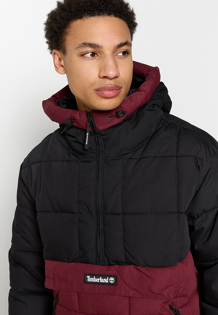 Czarna i bordowa kurtka puchowa z pikowanym wzorem, regulowanym kapturem i zapięciem na suwak z przodu. Posiada logo Timberland na klatce piersiowej.
